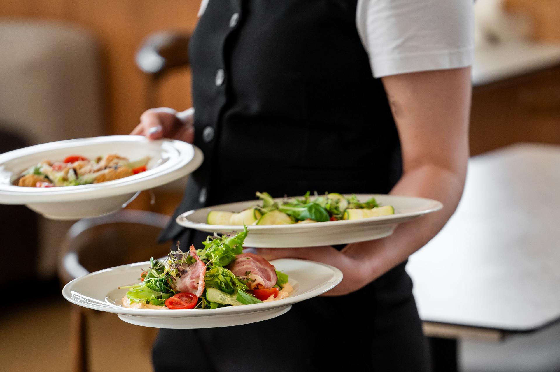 A waiter in a black uniform holds three plates with fresh salads, featuring greens, tomatoes, and ham, in a restaurant setting. The image captures professional service and dining presentation